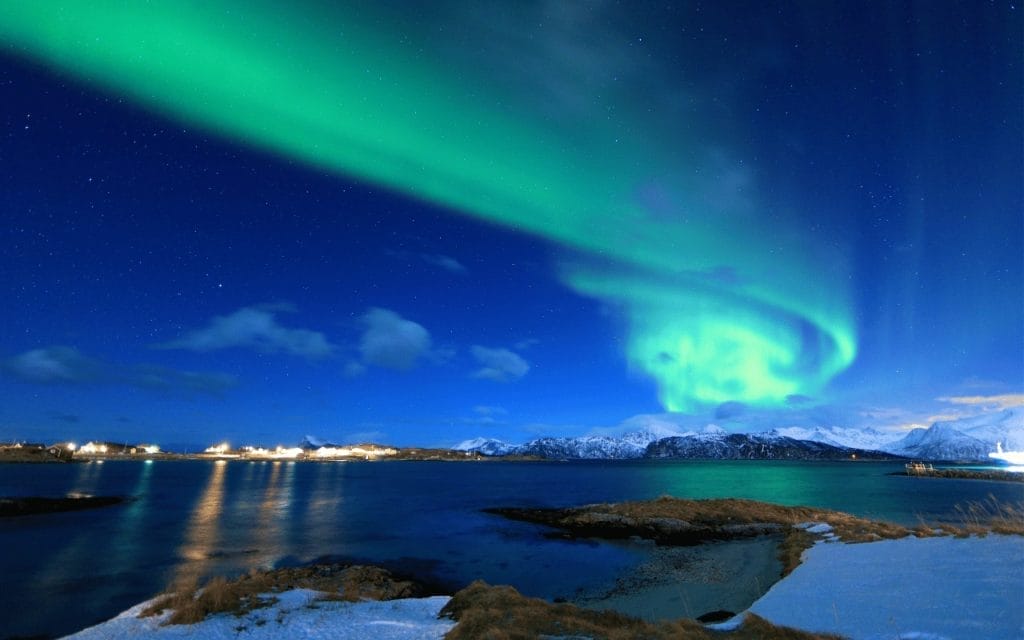 Le spectacle époustouflant des aurores boréales en Scandinavie au mois de janvier. Une aurore verte éclatante tourbillonne au-dessus du paysage enneigé, le bleu sombre du ciel nocturne parsemé d'étoiles et les lumières lointaines d'une colonie se reflétant sur les eaux glacées en dessous.