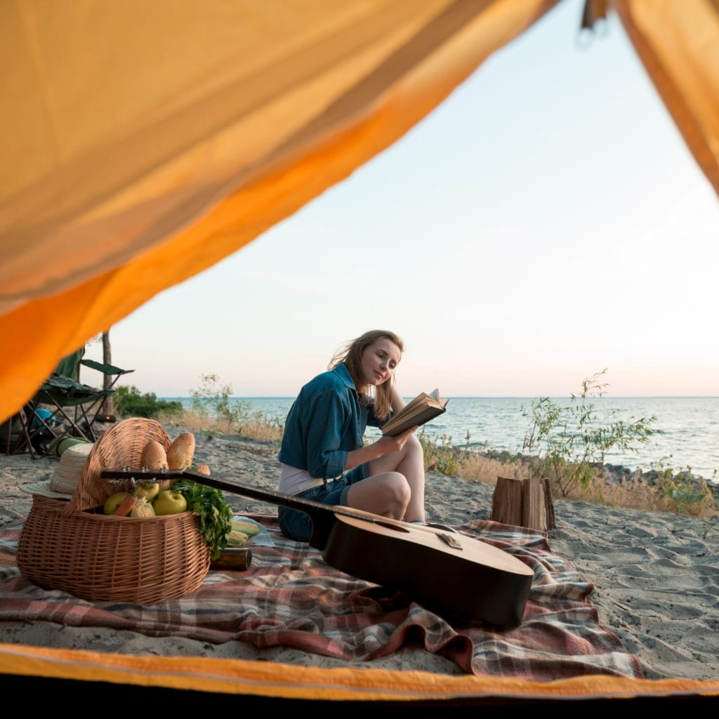 jeune femme lisant un livre près de son instrument de musique aux abords de sa tente sur la plage en bord de mer