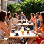 Terrasse de café à Pristina avec des personnes profitant de leurs boissons, dans une ambiance animée sous un soleil chaud, entourée de verdure.
