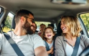 Une famille en voyage en voiture, les parents à l'avant et les enfants riant à l'arrière. Une journée ensoleillée avec un paysage verdoyant visible par les fenêtres de la voiture, capturant des moments de joie et de convivialité.