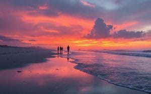 Vue de la plage d'Ostende pendant le coucher de soleil, avec un ciel vibrant aux couleurs orange et rose, des vagues douces et des silhouettes de personnes admirant le paysage.